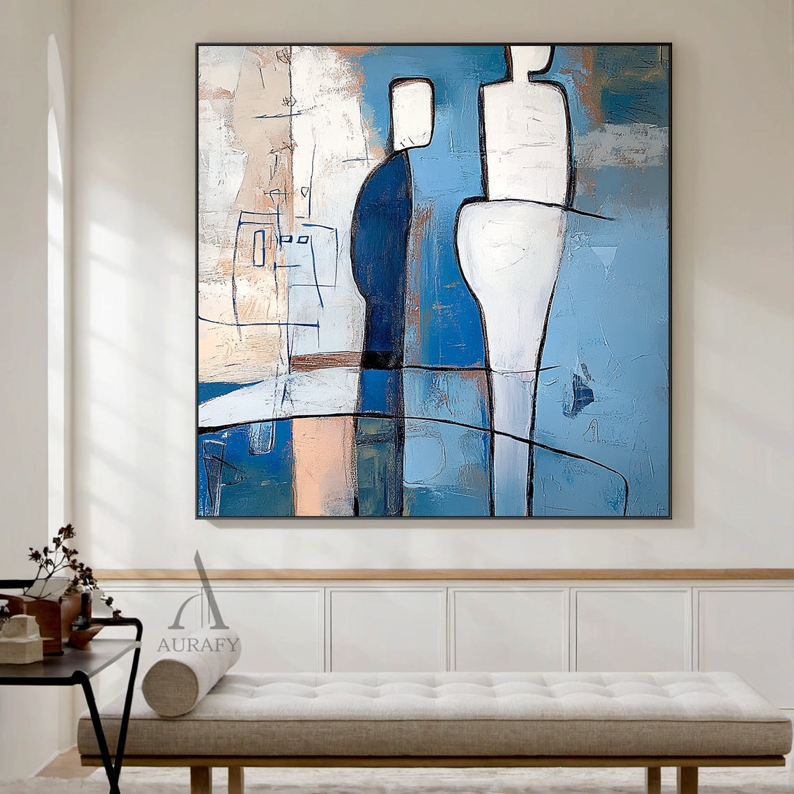Blue abstract wall art (AURA648) displayed hung on a warm white living room wall above a low-profile wooden console, featuring its signature tactile layered brushstrokes and bold black outlined geometric humanoid forms. The artwork includes two central contrasting figures: a deep navy blocky figure with white head, a taller fluid curved white figure, plus a delicate thin blue line sketch humanoid on the left, with a painterly background in sky blue, azure, navy tones paired with subtle warm beige and cream