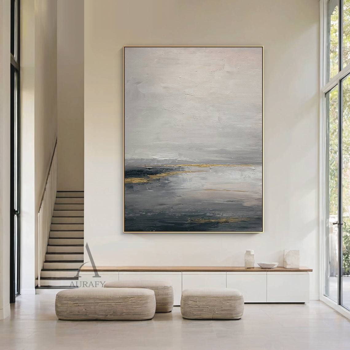 General Abstract gray beige canvas wall art (AURA626) displayed in modern home interior scene, features the signature layered muted gray shades (light, medium, dark) base with soft beige transition zones, accented with subtle shimmering golden streaks and patches. The tactile textured brush stroke layers present well against the neutral backdrop: loose hazy upper light gray and beige areas evoke misty cloud imagery, thick deep gray lower sections mimic dark water surface, golden elements simulate natural li