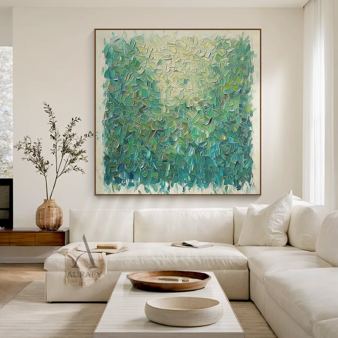 Semi-abstract impressionist green landscape impasto canvas painting, handcrafted with thick palette knife short textured strokes. This displayed hanging piece features a signature natural tone gradient, shifting from pale yellow and soft sunlit green on the upper half down to grass green, olive green and dark teal green on the lower half to recreate layered shaded foliage texture, with a light beige raw canvas border framing the whole artwork. It is presented against a muted off-white wall paired with light