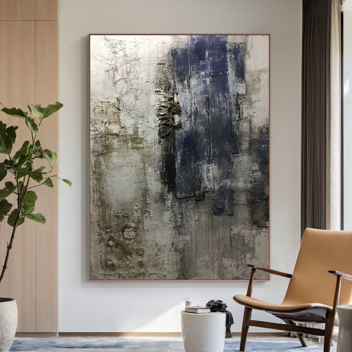 AURA223 Wabi Sabi Abstract Wall Art with layered impasto strokes and cracked textures in brown, black, deep blue, and neutral tones. Displayed in a modern living room setting with beige armchair, wooden cabinet, green plant, and blue rug—ideal for living room, bedroom, or office decor. Embodies Wabi Sabi philosophy of beauty in imperfection, adding depth to minimalist, rustic, or modern spaces. Tactile organic finish makes it a focal point to elevate home ambiance. Unique handcrafted abstract painting for t