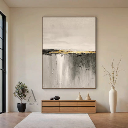 Hand-painted beige abstract impasto canvas wall art AURA718, displayed hanging on a matte warm white living room wall above a light oak low sideboard in a real neutral home styling scene, the artwork features soft warm off-white and light beige with subtle thick paint grain texture on the upper section, central horizontal matte charcoal stripe accented with raised grainy metallic gold thick paint band as focal point, lower area is gradient gray from light beige gray to dark charcoal with vertical sweeping b