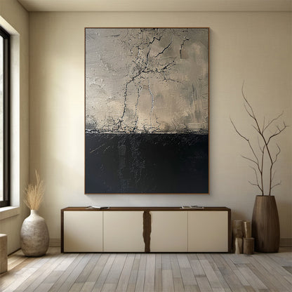 Vertical abstract painting with upper beige-gray section (crack-like linear patterns, impasto texture) and lower deep black section, framed in thin warm brown. Hung above modern light beige console table with dark brown accents in a minimalist room: soft beige walls, light gray wood plank flooring, light beige ceramic vase (dried grass) and dark brown wooden vase (bare branches) nearby. This piece serves as a focal point in the serene, sophisticated space.