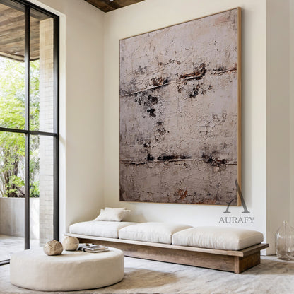 Large vertical abstract wall art with a textured, cracked surface in neutral beige, gray, and subtle brown/black tones, framed in a thin brown border. Displayed in a sleek modern living room setting above a low-profile linen sofa, adjacent to a minimalist black metal floor lamp and a small marble decorative tray holding a citrus-scented candle—ideal for infusing contemporary living spaces with understated artistic charm.