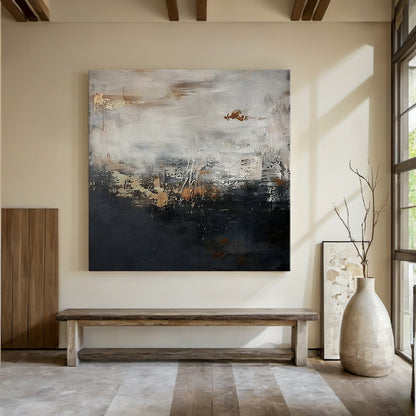 Wabi Sabi abstract sculpted canvas art with impasto knife strokes and tactile surface—deep black lower section, light gray upper layer, golden copper accents. Displayed in a serene modern organic space: light beige walls, wooden ceiling, soft-cushioned bench, gray rug, metallic vase with branches, natural light. Embraces Wabi Sabi’s imperfect beauty and subtle warm softness (Pink vibe) for calm, minimalist home decor. AURA196.