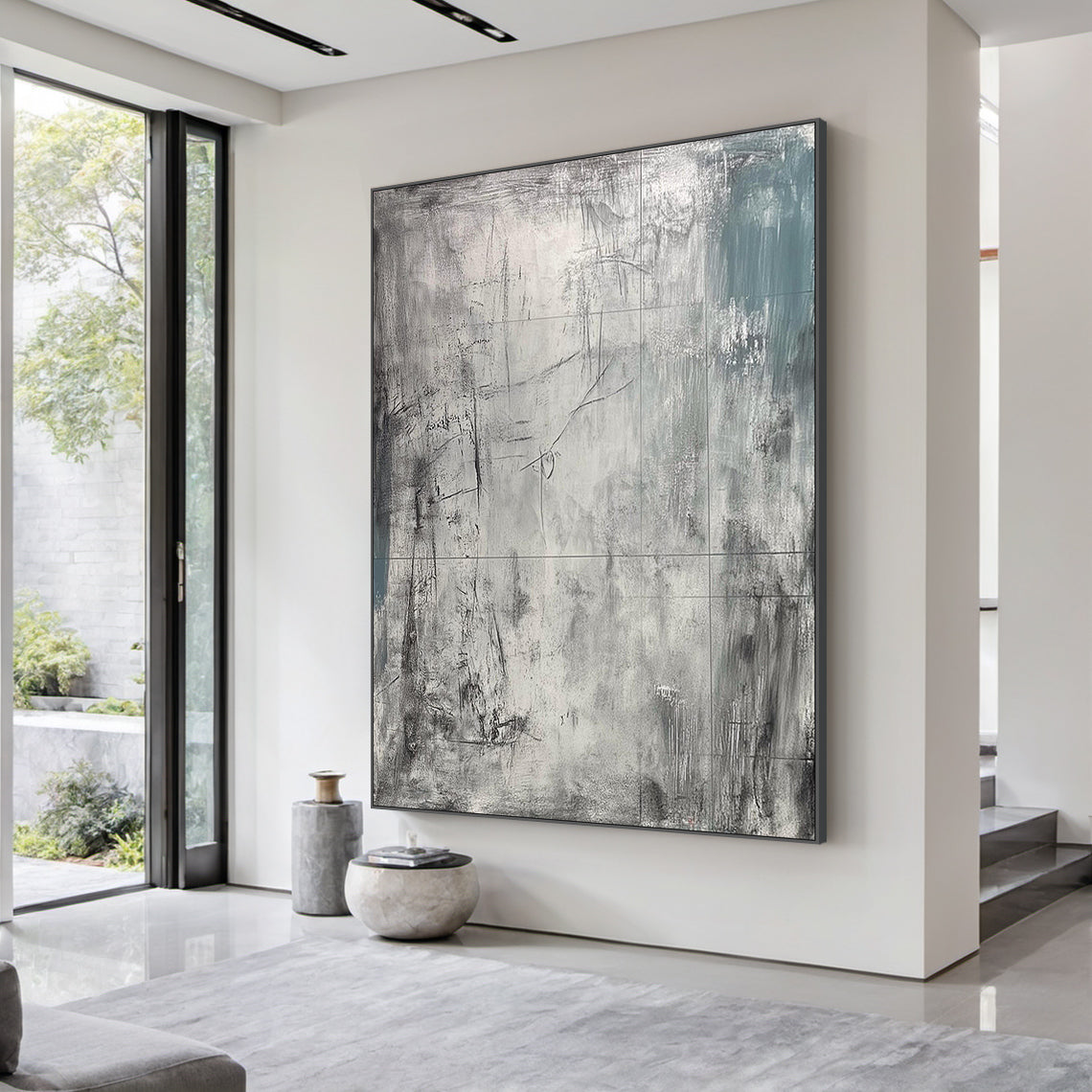 AURA224 Wabi Sabi Gray Abstract Textured Canvas Wall Art - Large statement piece with layered tactile brushstrokes, muted grays, and subtle teal accents, displayed above a plush beige linen sofa in a neutral-toned modern living room. Soft warm light from a wooden floor lamp illuminates the space, complementing a minimalist marble coffee table with a ceramic vase of dried pampas grass and sleek metal side chairs. Ideal focal point for calm modern living spaces, adding organic tactile depth to neutral decor p