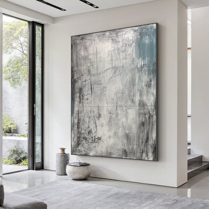 AURA224 Wabi Sabi Gray Abstract Textured Canvas Wall Art - Large statement piece with layered tactile brushstrokes, muted grays, and subtle teal accents, displayed above a plush beige linen sofa in a neutral-toned modern living room. Soft warm light from a wooden floor lamp illuminates the space, complementing a minimalist marble coffee table with a ceramic vase of dried pampas grass and sleek metal side chairs. Ideal focal point for calm modern living spaces, adding organic tactile depth to neutral decor p
