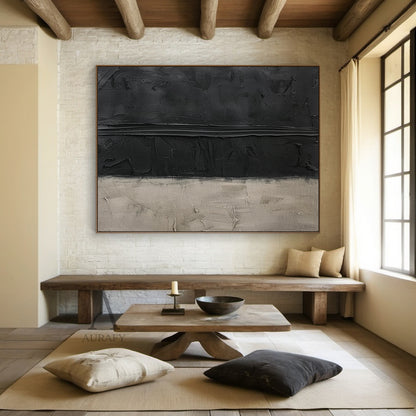 Handcrafted wabi-sabi Japandi abstract canvas artwork (AURA797) wall-mounted scene display, featuring a two-tone muted neutral palette of deep matte black and soft warm beige. The piece uses thick hand-applied impasto technique: upper black areas have scored, scraped pigment textures with a prominent raised horizontal impasto stripe across the middle, lower beige section has plaster-like aged, scratched, faded uneven surfaces. Minimalist textured abstract wall art presented in real home space, suitable for 