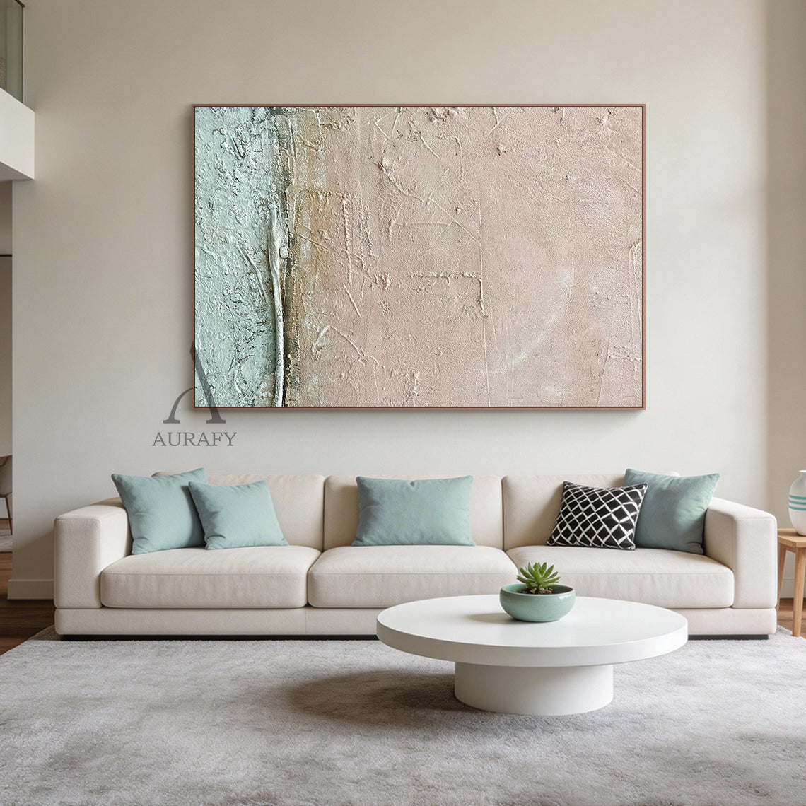 Wabi Sabi abstract wall art with textured impasto finish—soft blush pink upper layer, light teal green lower section, and neutral beige transitions—framed in thin brown wood. Displayed in a minimalist living room with warm taupe walls, walnut coffee table, neutral linen sofa, woven jute rug, and a small ceramic vase holding dried pampas grass. AURAFY modern home decor piece ideal for adding serene, imperfect beauty to cozy contemporary spaces.